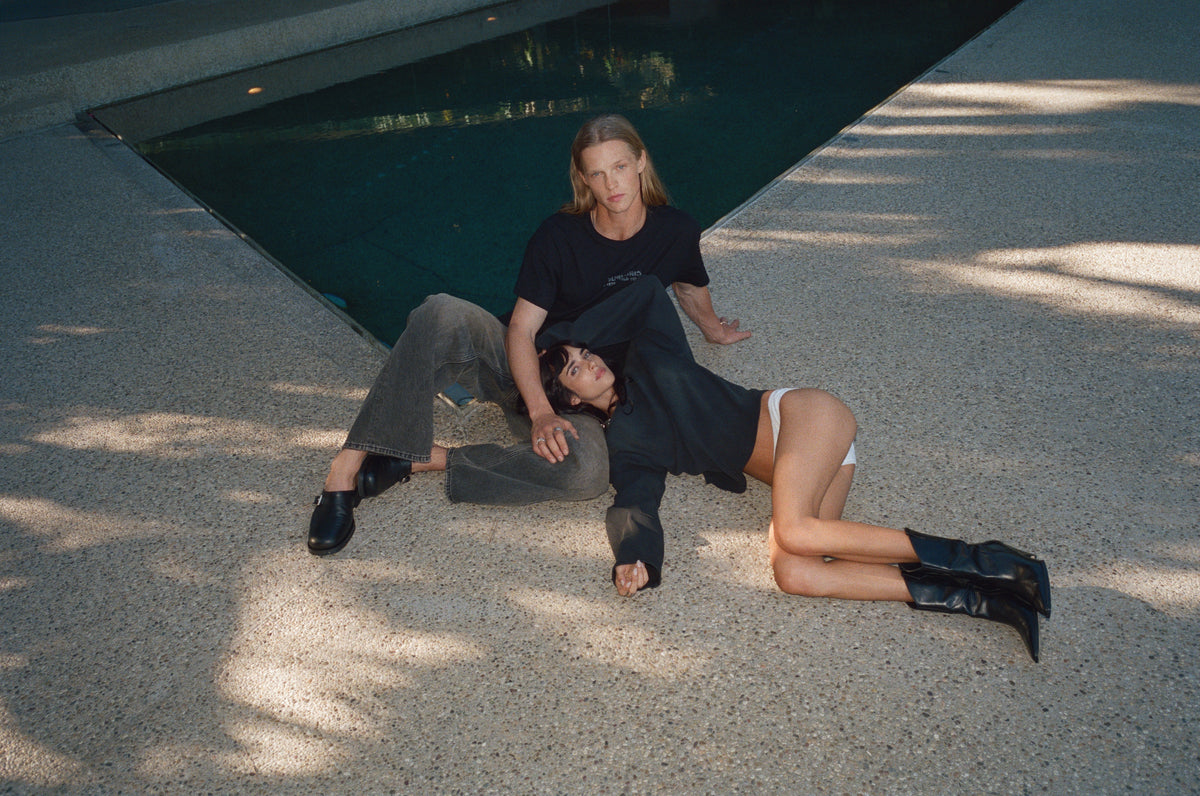 Two models in black sitting on a sunlit terrace beside the pool.