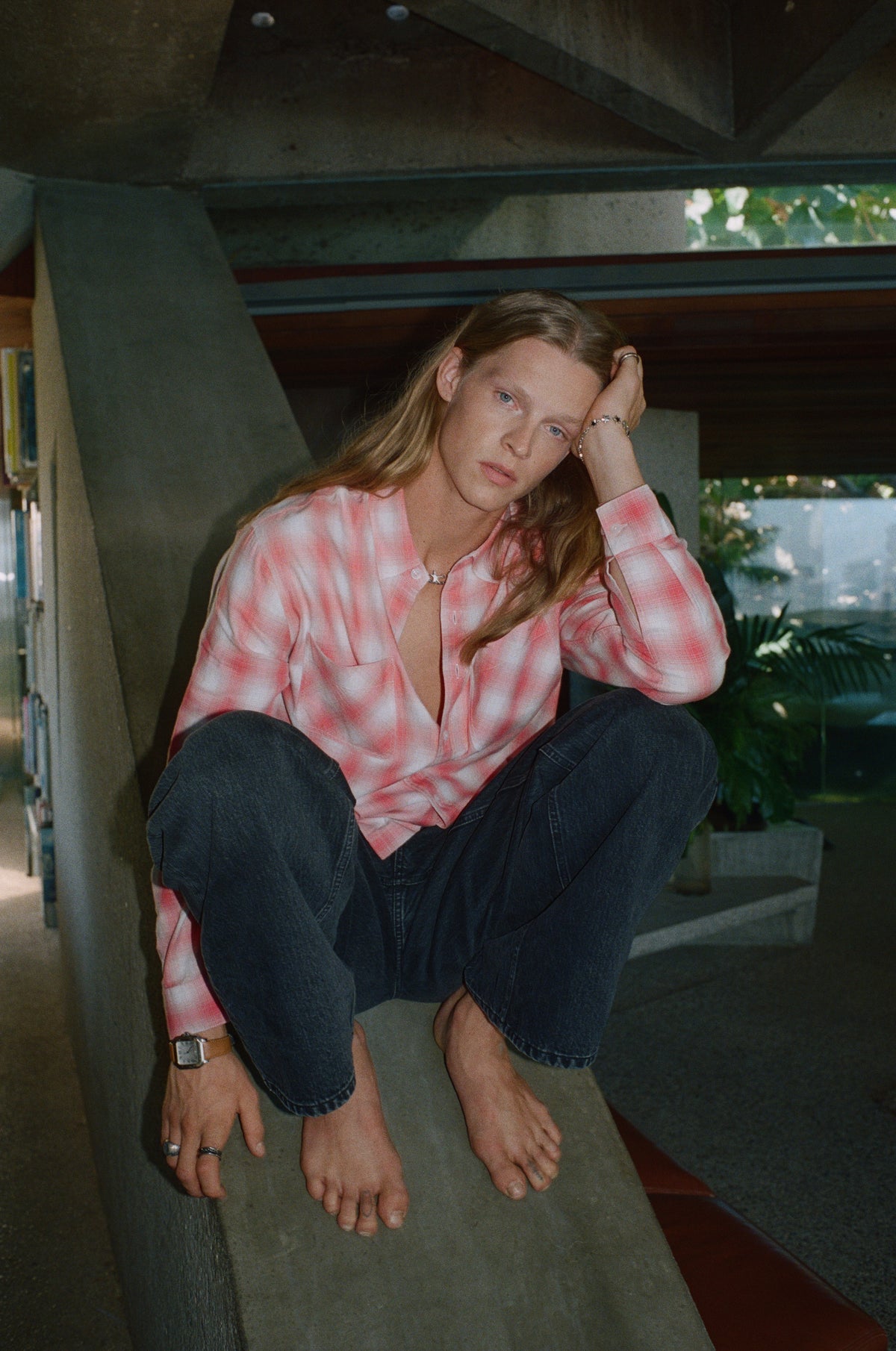 Model in a pink checked shirt and dark jeans sitting beneath an angular concrete roof.