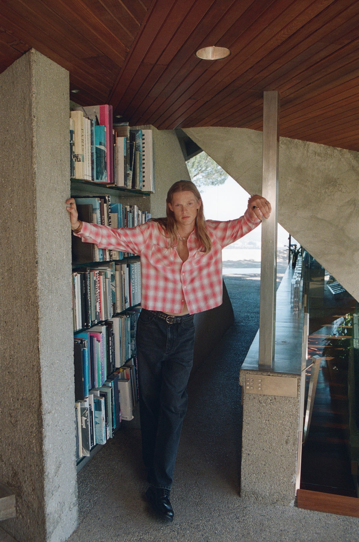 Model in a pink checked shirt and dark trousers standing beside bookshelves.