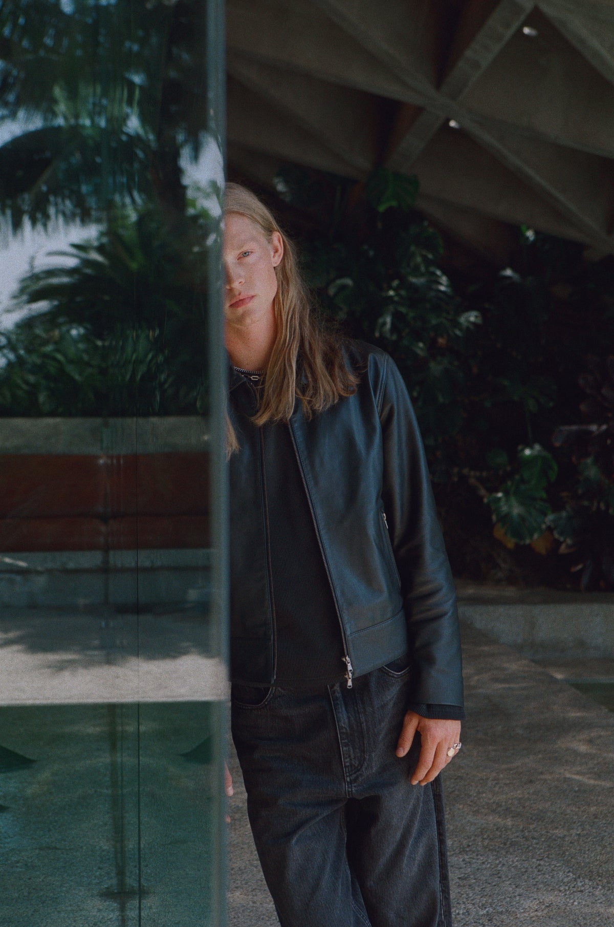Model in dark denim standing beside a reflective glass wall.