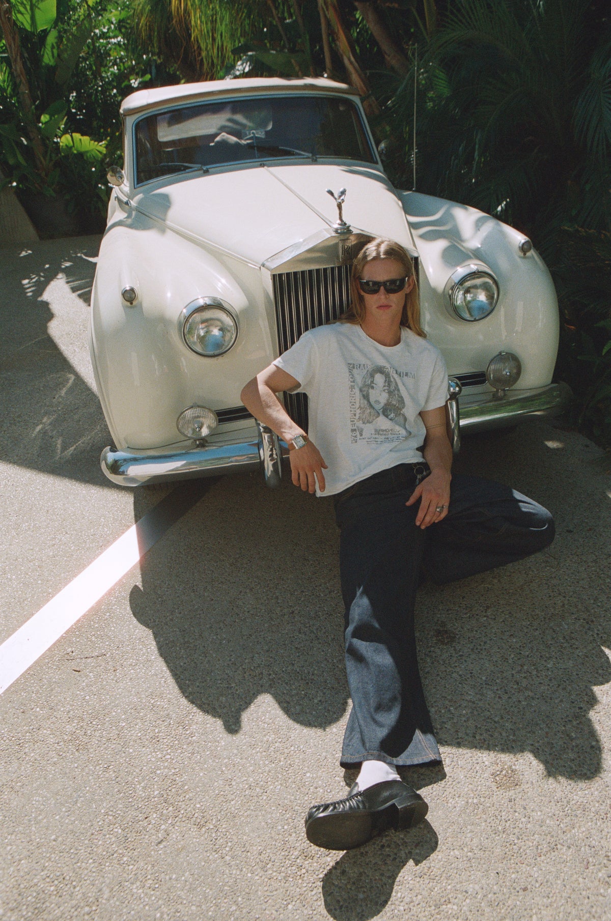Model in a white graphic T-shirt and dark jeans sitting in front of a white vintage car.