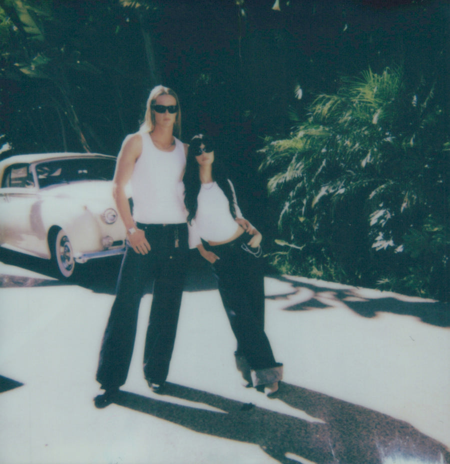 Campaign polaroid: two models in white tanks and dark trousers on a driveway beside a white vintage car