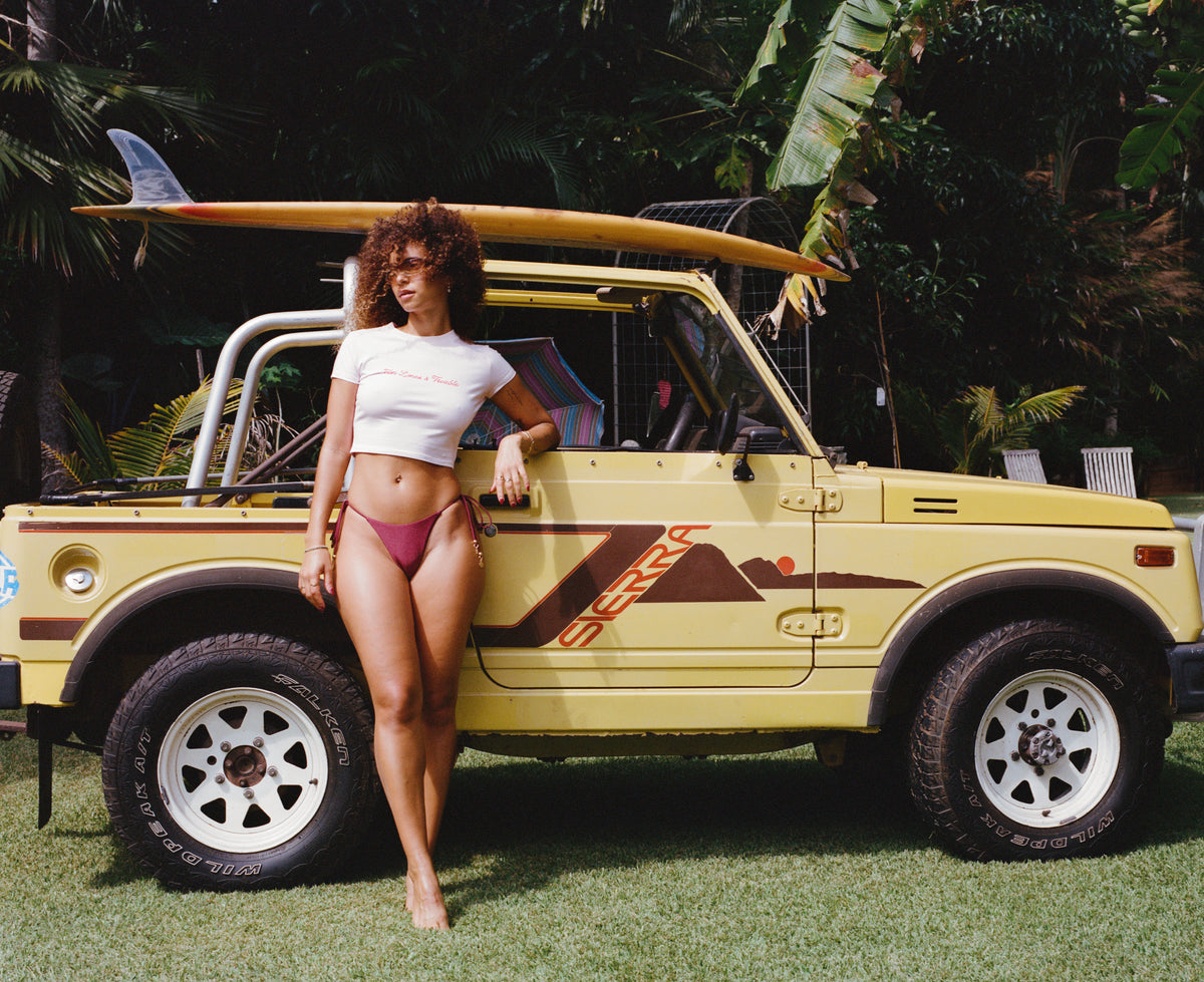 Model in a white cropped T-shirt and burgundy bikini bottoms leaning against a yellow four-wheel drive with surfboards on its roof.