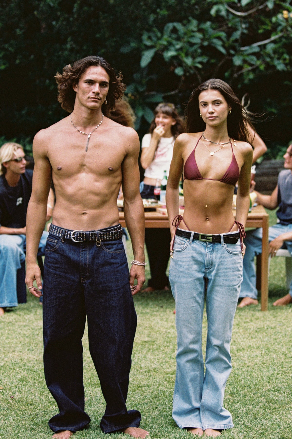 Two models in wide-leg jeans walking across a lawn, one wearing a burgundy bikini top.