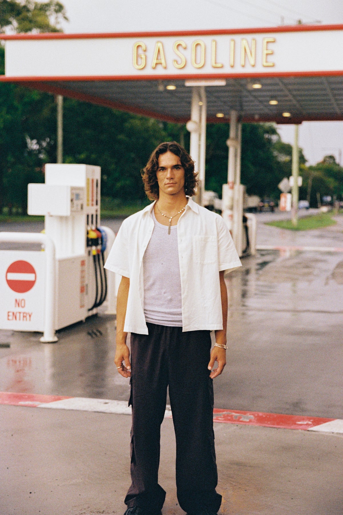 Model in an open white shirt and black trousers beneath a gasoline sign at a wet petrol station.