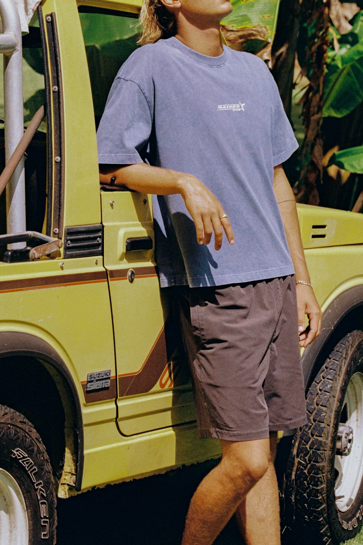 Cropped view of a model in a purple T-shirt and brown shorts beside a yellow four-wheel drive.