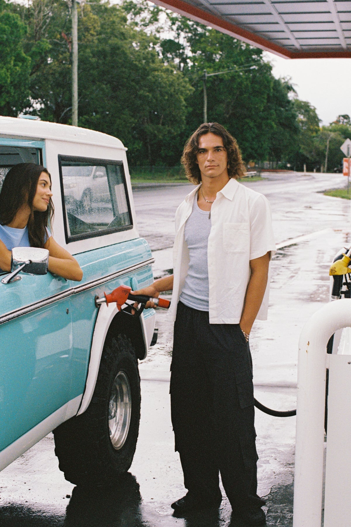 Model in an open white shirt and black trousers refuelling a turquoise four-wheel drive, with another model leaning from the window.