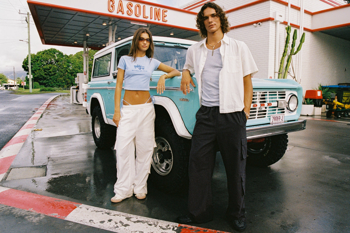 Wide view of two models in a blue cropped T-shirt and an open white shirt beside a turquoise four-wheel drive at a petrol station.