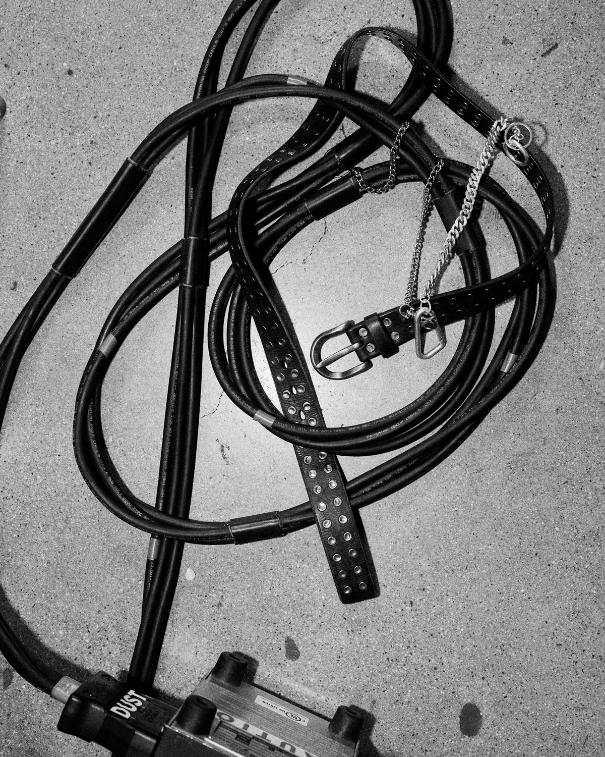 A studded leather belt coiled with reins on concrete, photographed in black and white.