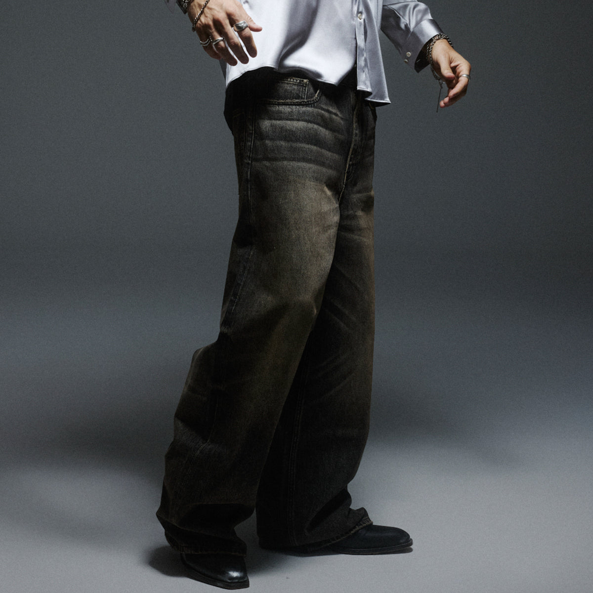A model wearing faded dark wide-leg jeans and a pale satin shirt against a grey studio backdrop.