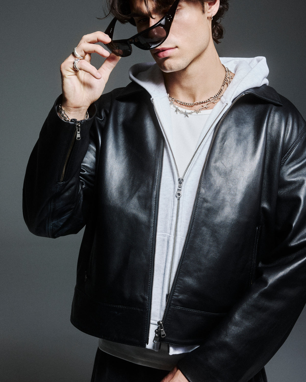 A model in a black leather jacket over a grey hoodie, lifting their sunglasses.