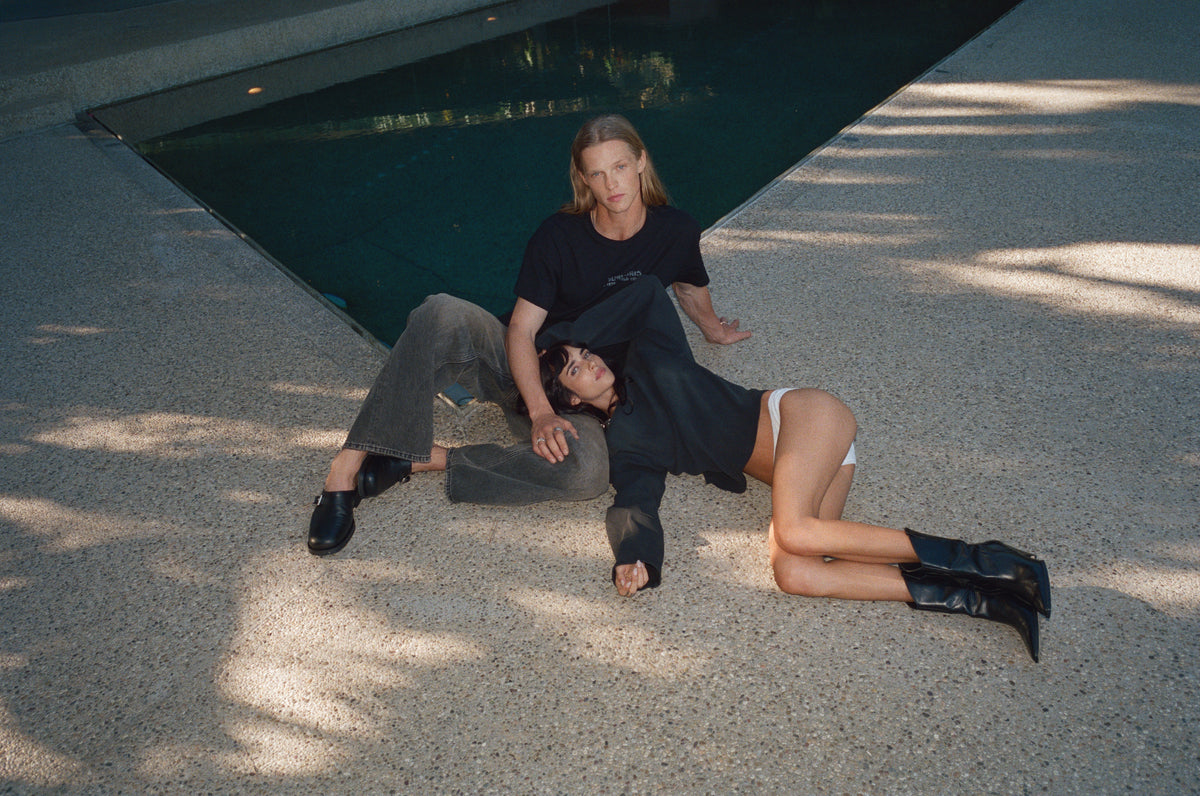 Two models lying on the concrete beside a pool, in a black T-shirt, jeans and boots.