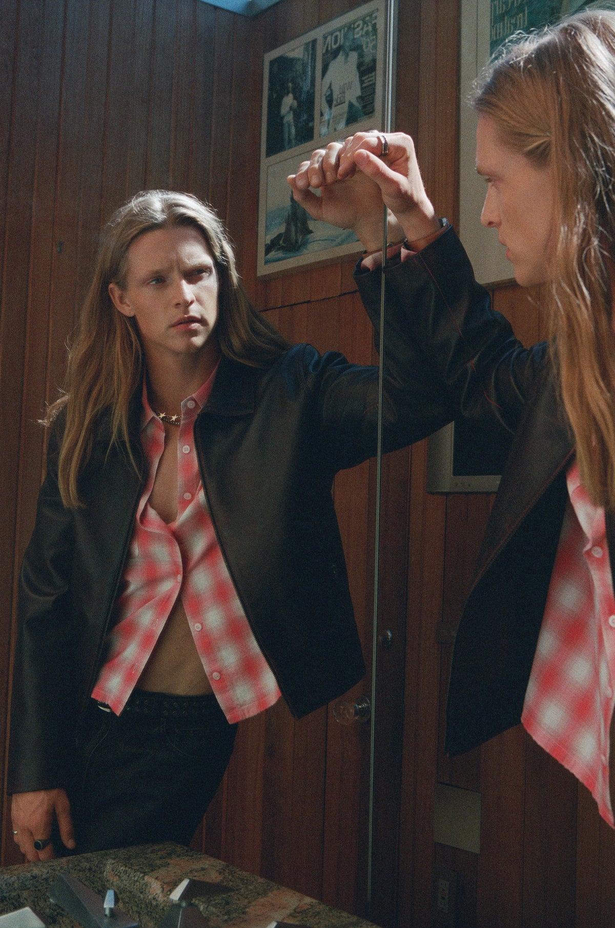 Model in a pink checked shirt and dark jacket reflected in a mirror.