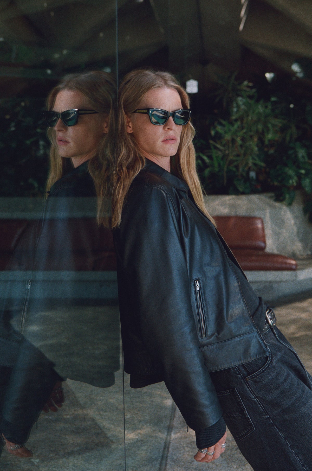 Model in a dark leather jacket and sunglasses reflected in glass.
