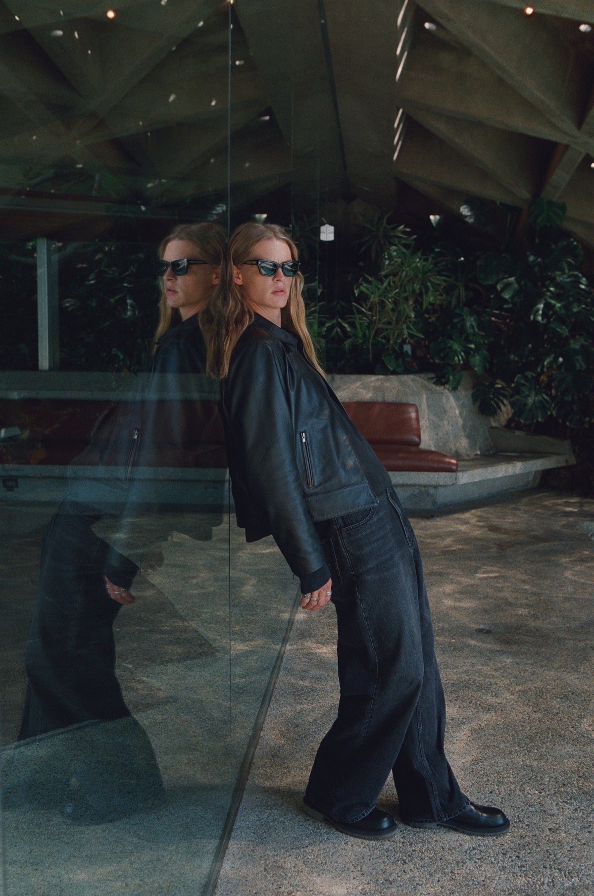 Full-length view of a model in dark clothing leaning beside reflective glass.