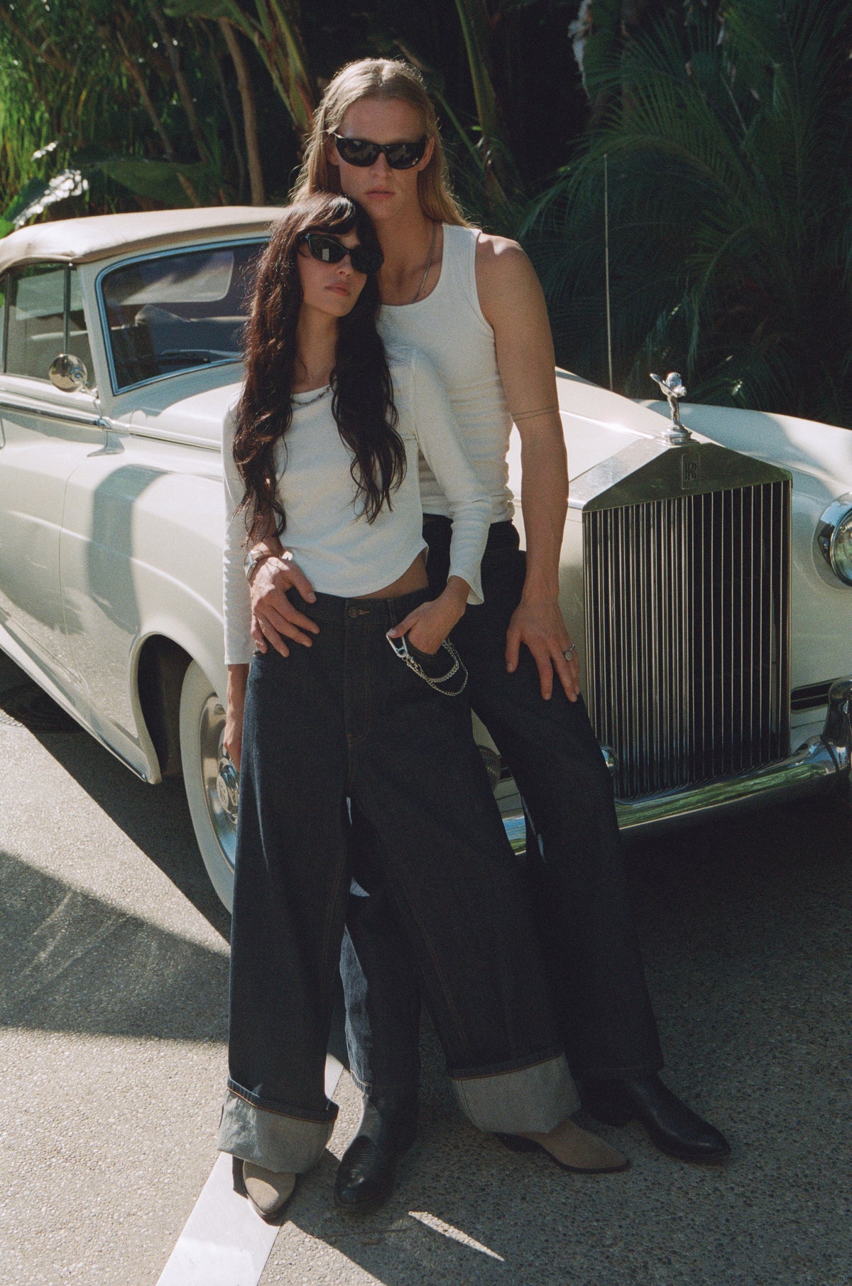 Two models in white tops and dark trousers beside a white vintage car.