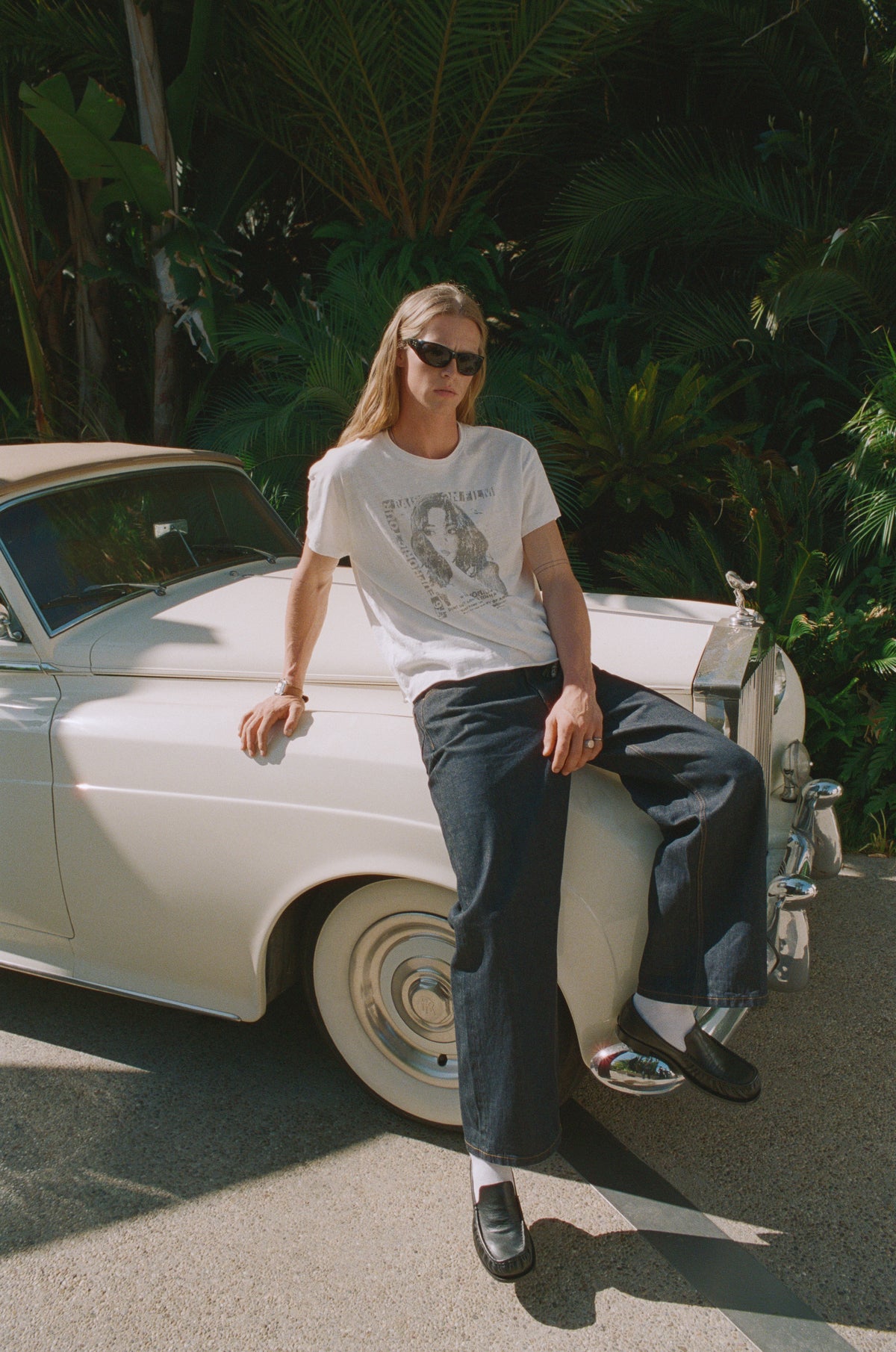 Model in a graphic T-shirt and dark jeans sitting on a white vintage car.