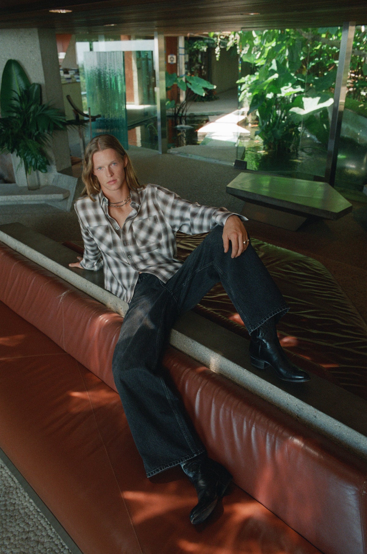 Model in a checked shirt and dark trousers reclining on an orange leather sofa.