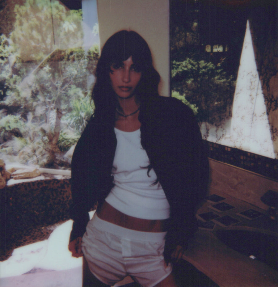 Campaign polaroid: a model in a white tank and open dark shirt outdoors beside a pale wall