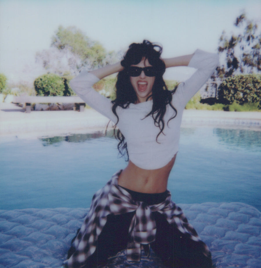 Campaign polaroid: a model at a pool edge in a white crop top and check flannel, arms raised