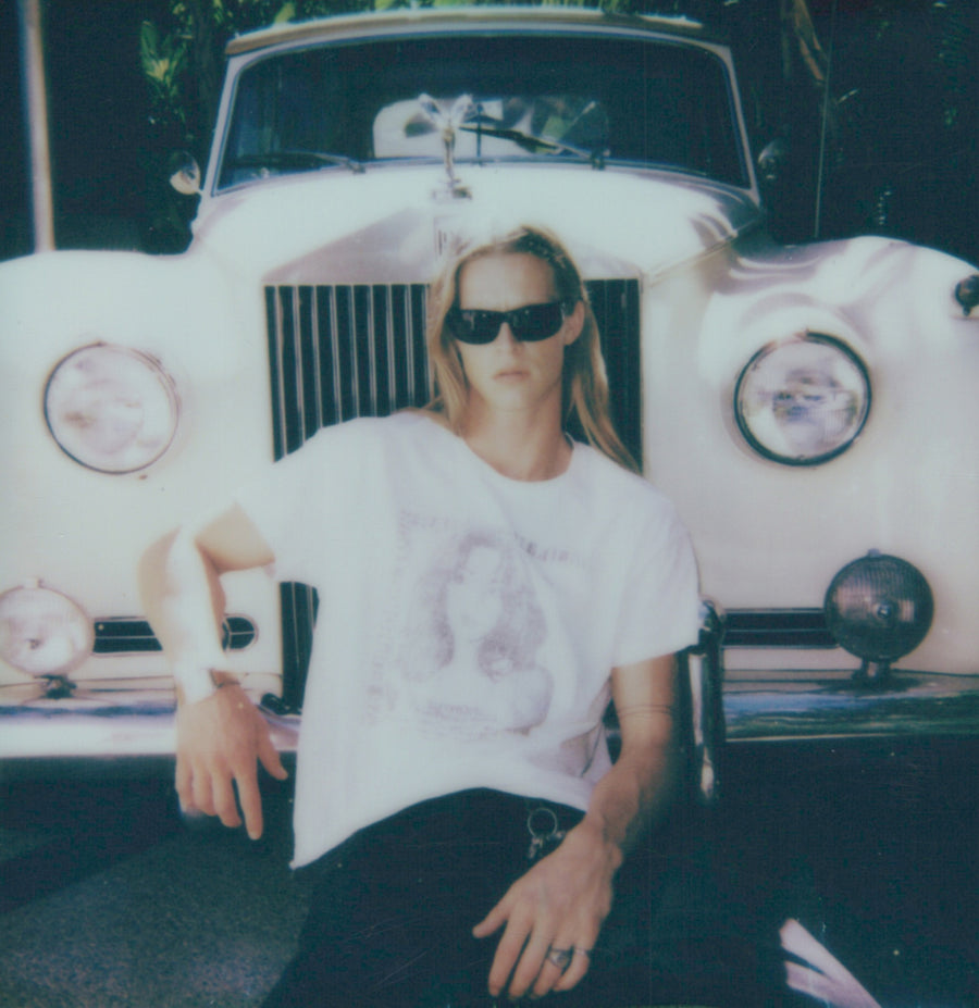 Campaign polaroid: a model in a printed white tee seated on the bumper of a white vintage car
