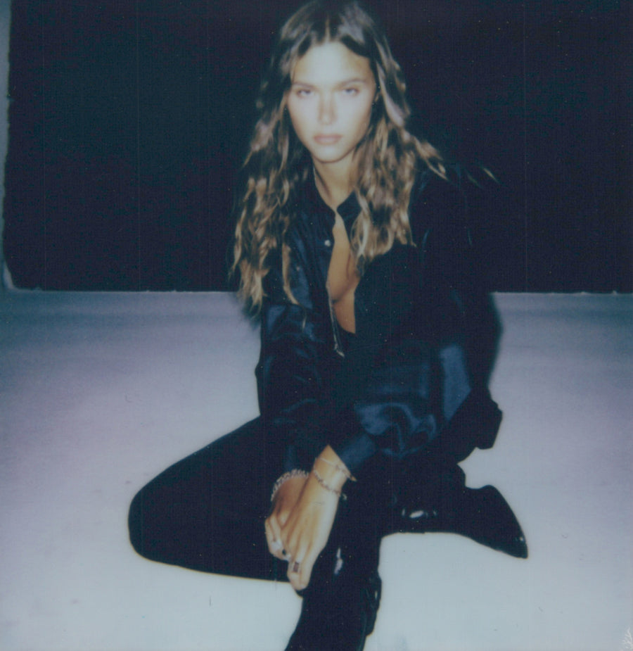 Campaign polaroid: a model in a black leather jacket seated on a dark studio floor