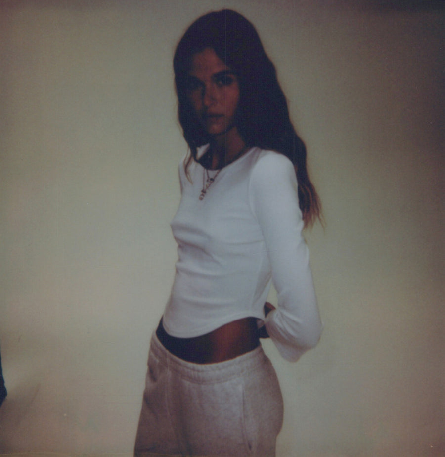 Campaign polaroid: a model in a white long-sleeve and pale trousers against a plain studio backdrop