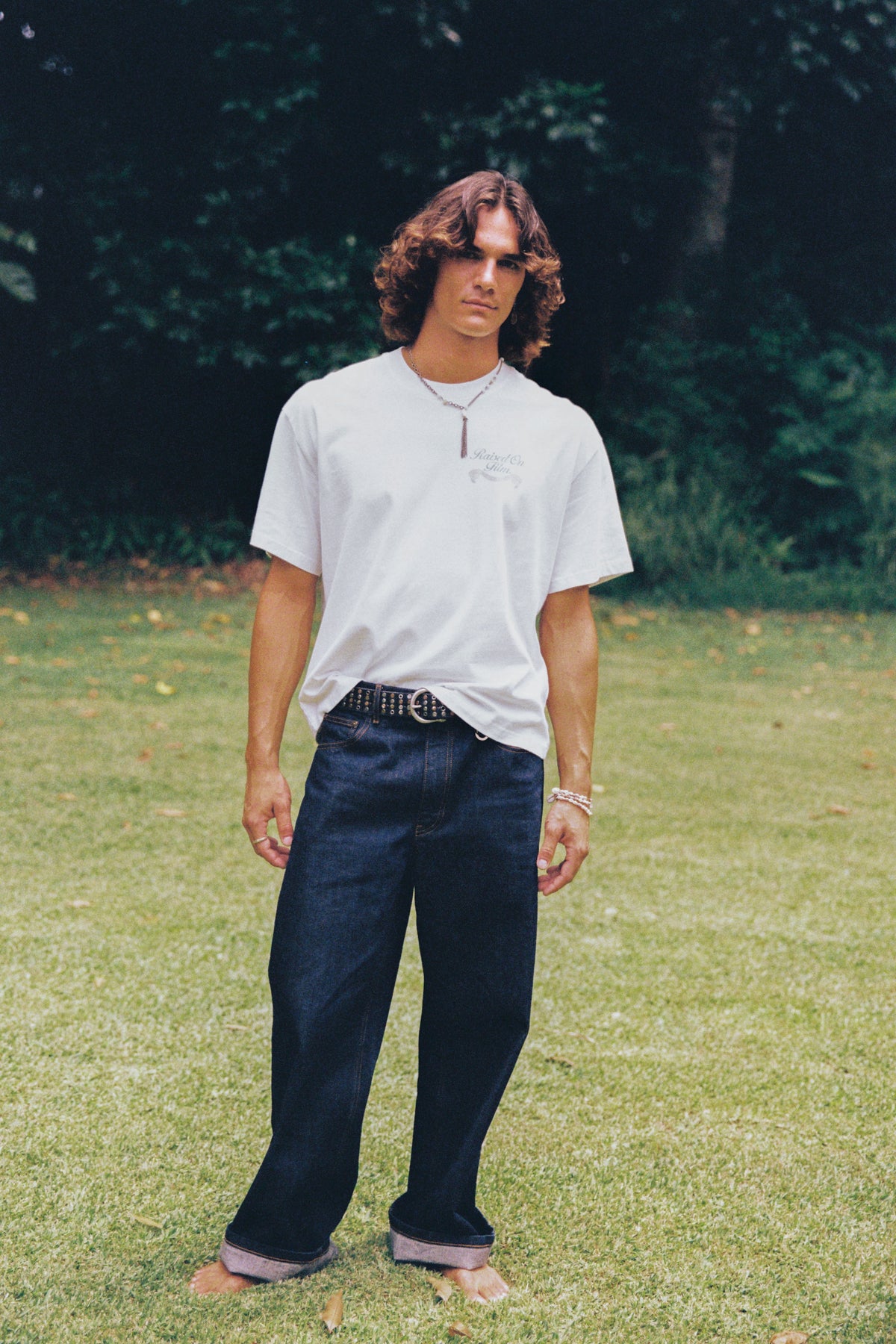 Model in a white T-shirt and dark wide-leg jeans standing barefoot on a lawn.