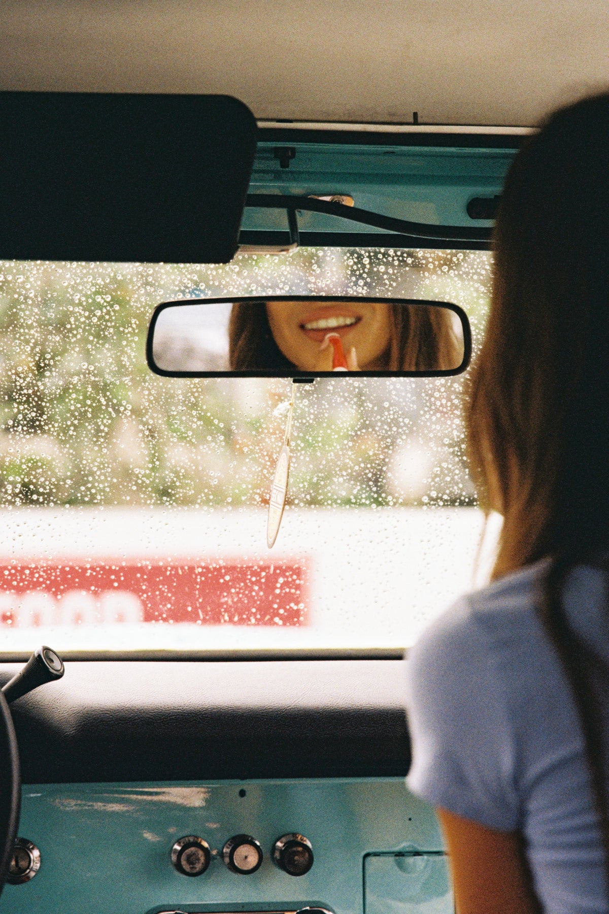 View from inside a turquoise four-wheel drive, with a model in a blue T-shirt reflected in the rear-view mirror and rain on the windscreen.